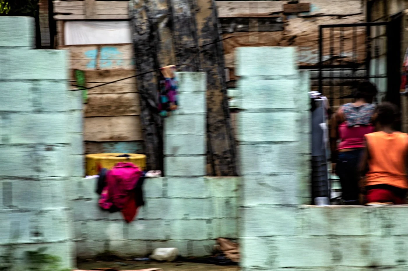 People near a wall made of light green concrete blocks, with some wooden panels and black metal fencing, in an outdoor setting.