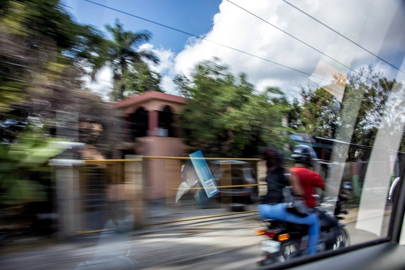 Blurry photo of a street scene taken from a moving vehicle, showing a person riding a motorcycle with a passenger, houses with trees, and overhead power lines under a partly cloudy sky.