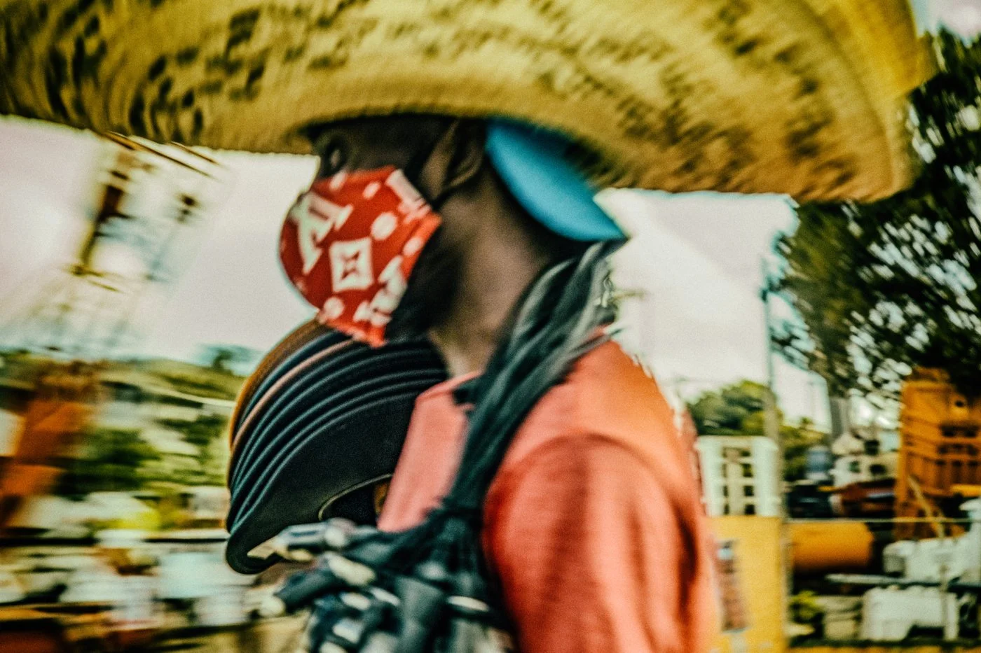 Person wearing a patterned bandana mask, carrying a guitar, and wearing an orange jacket, walking outdoors with blurred trees and buildings in the background.