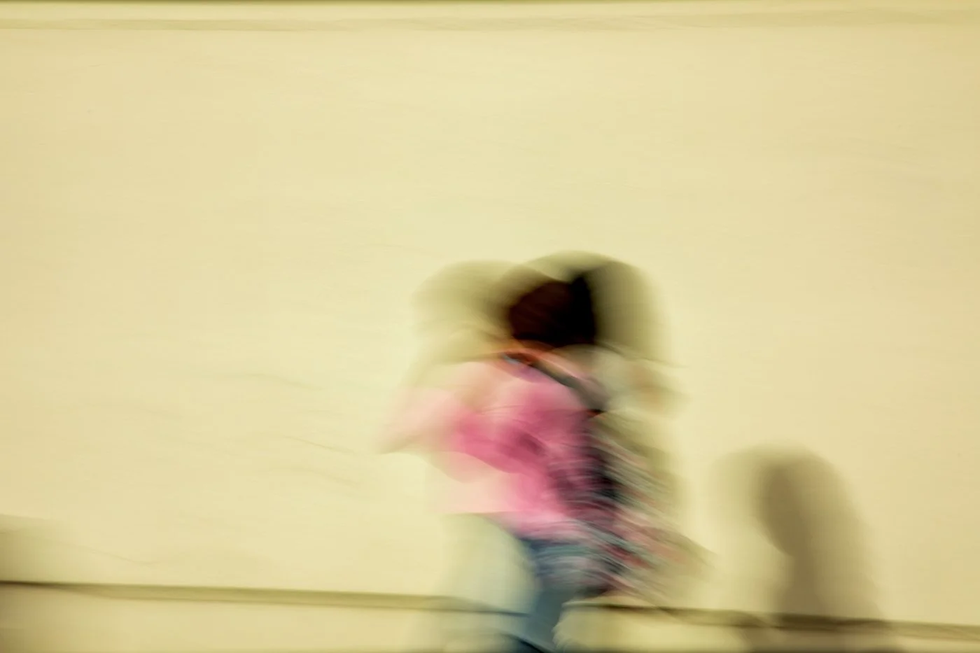 A person walking with a pink jacket against a plain light-colored wall, with a shadow on the floor.
