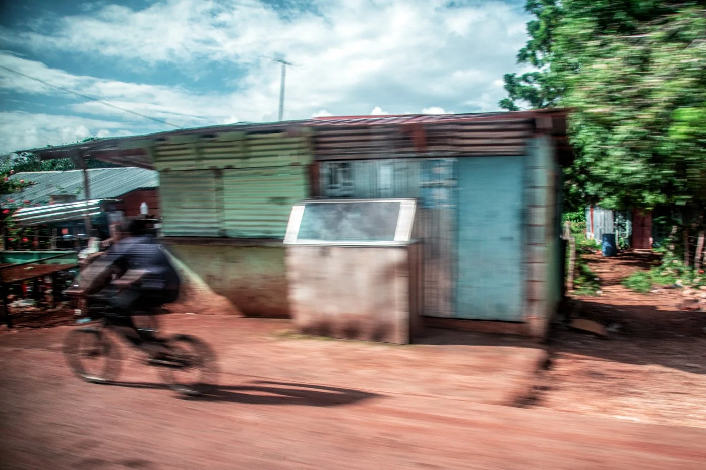 A blurred motion photo of a person riding a bicycle past a rustic building made of metal sheets in a rural area with dirt road and green trees.