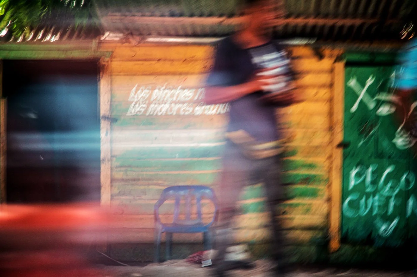 Blurred image of a person standing in front of a colorful wooden wall with some text, with a plastic chair in the foreground and a green door to the right.