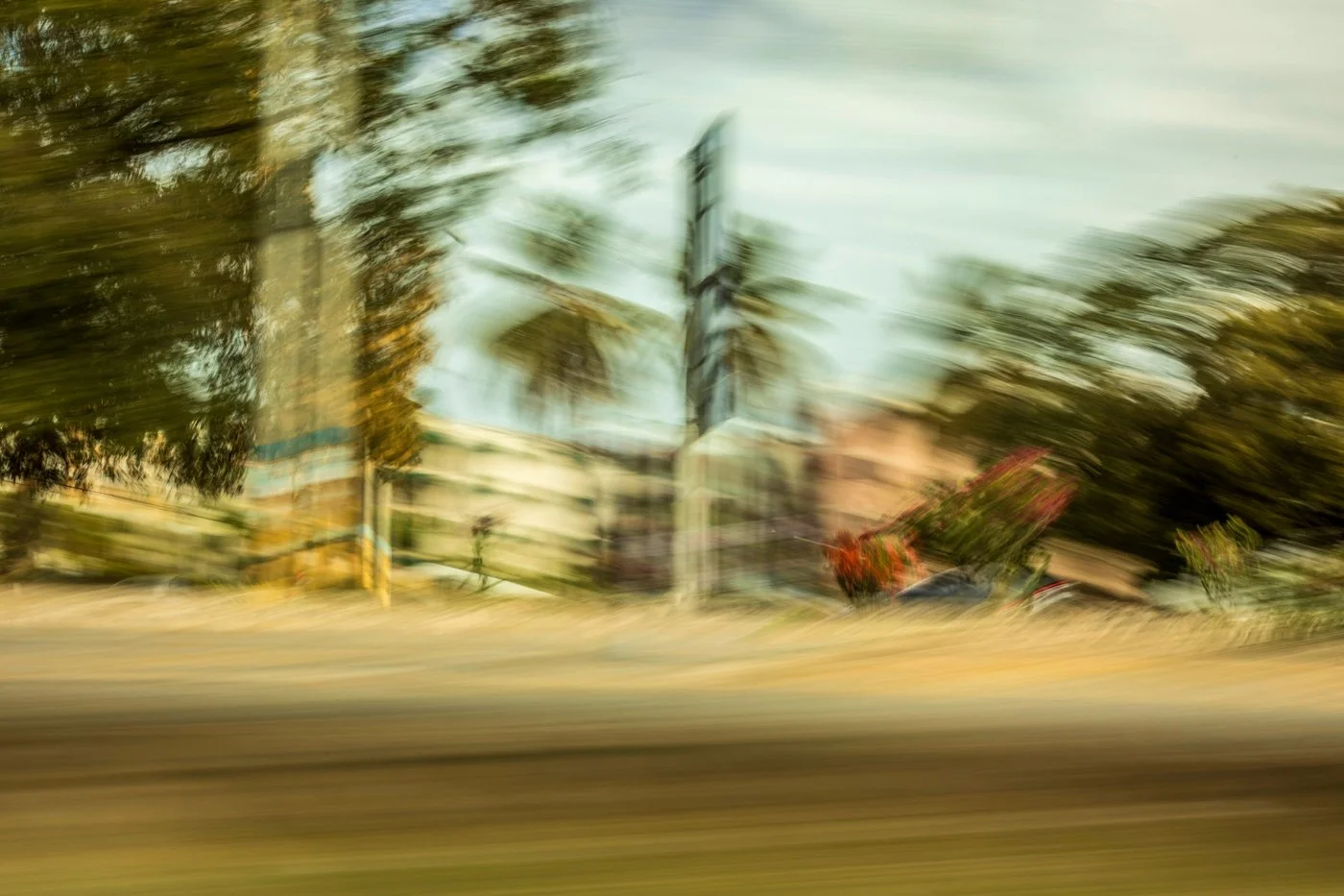 Motion-blurred image of a city street with trees, buildings, a billboard, and some greenery.