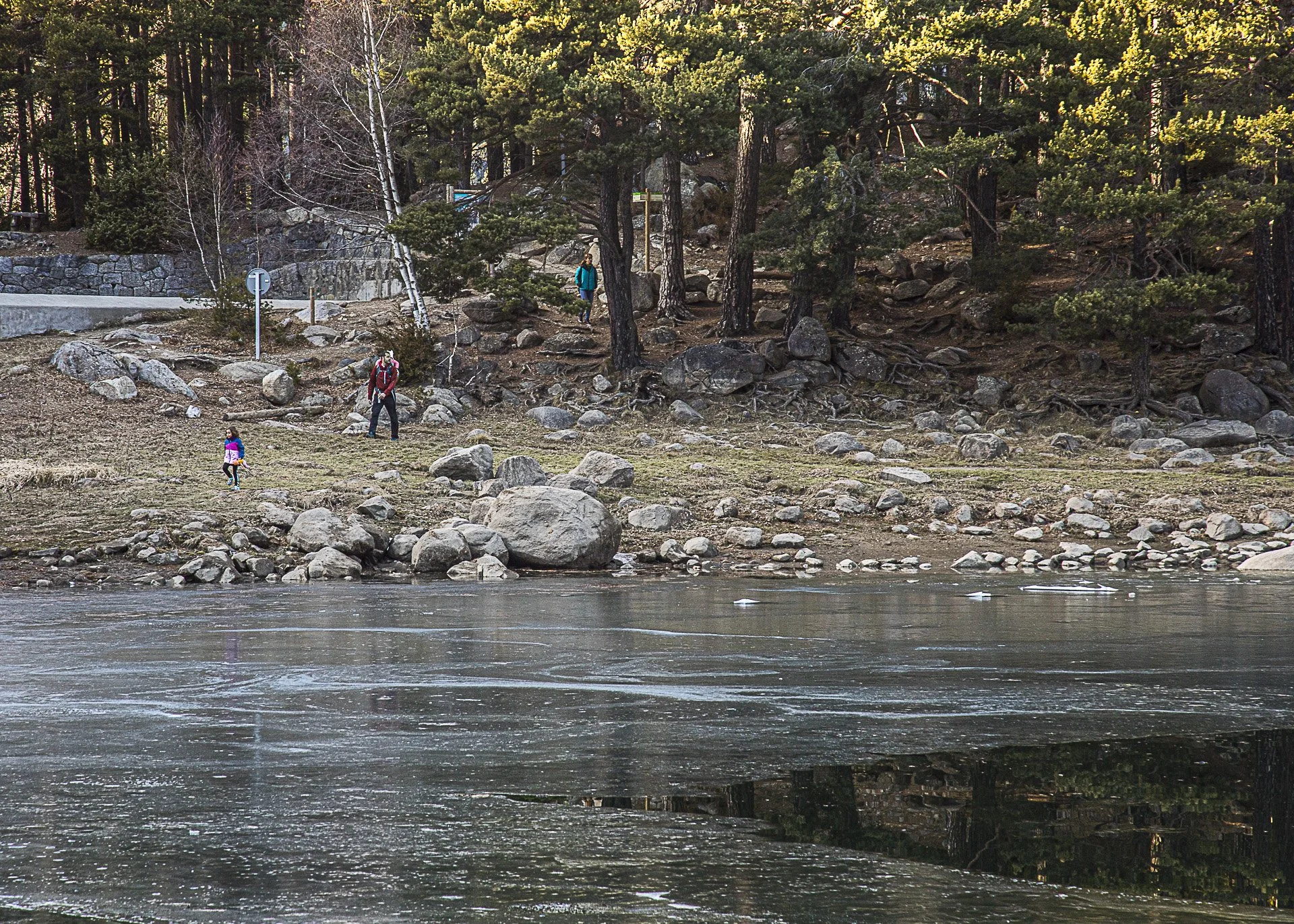 Three people walking along a rocky riverside surrounded by trees and forest, with a small girl wearing a pink jacket in the foreground.