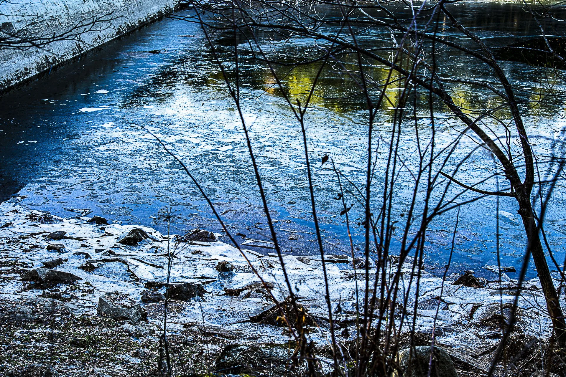 A partially frozen river with ice on surface, rocks along shore, and leafless branches in foreground.