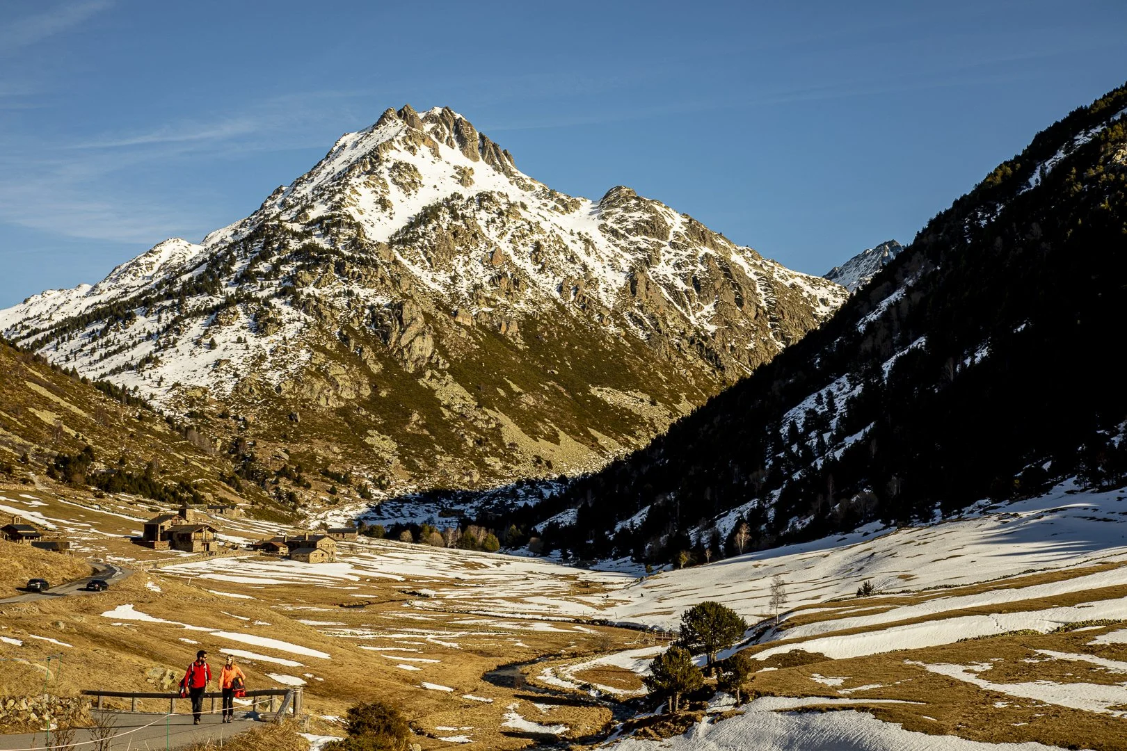 People hiking on a trail in a mountainous landscape with snow patches, green trees, and a village at the base of snow-covered peaks under a clear blue sky.