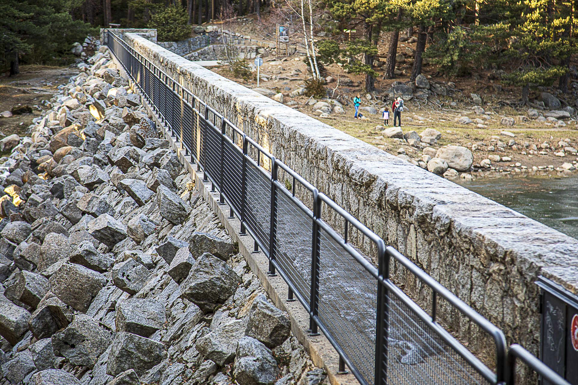 A long stone bridge with a black metal railing spans over a rocky bank beside a river, with a wooded hillside and three people walking in the distance.