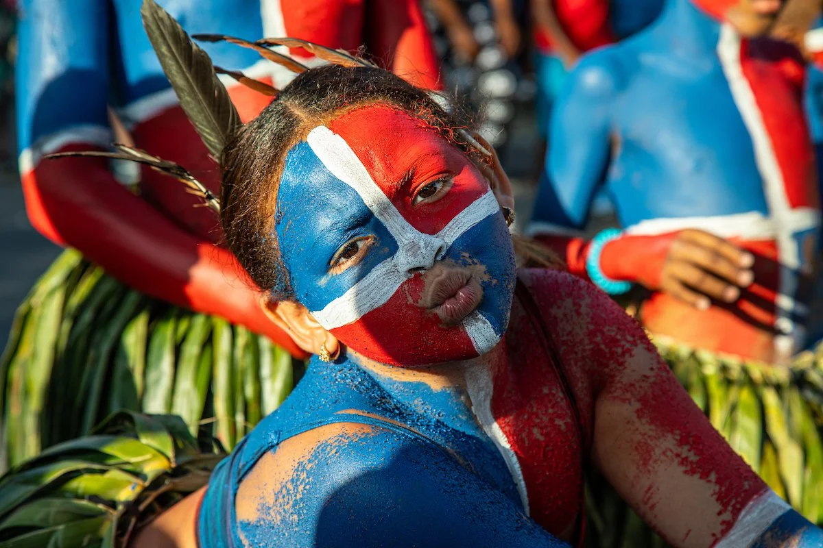 Girl with face painted with the flag of Norway, surrounded by people with similar face paint and colorful clothing, celebrating outdoors.