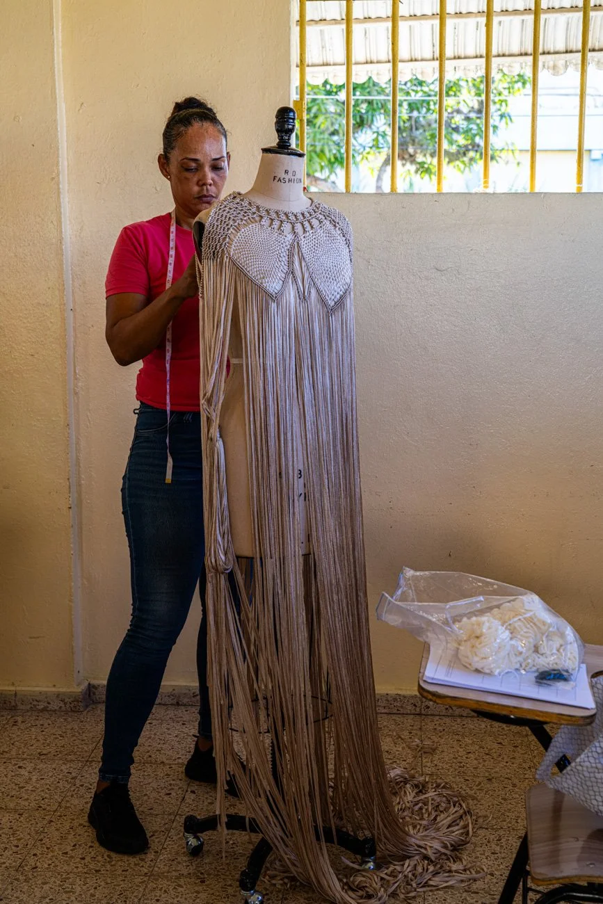 A woman working on a dress with long fringe details on a dress form in a room with cream-colored walls and a window with metal bars.
