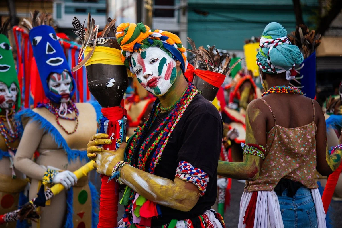 People dressed in colorful costumes and face paint participating in a cultural parade or festival, with some holding decorated staffs.
