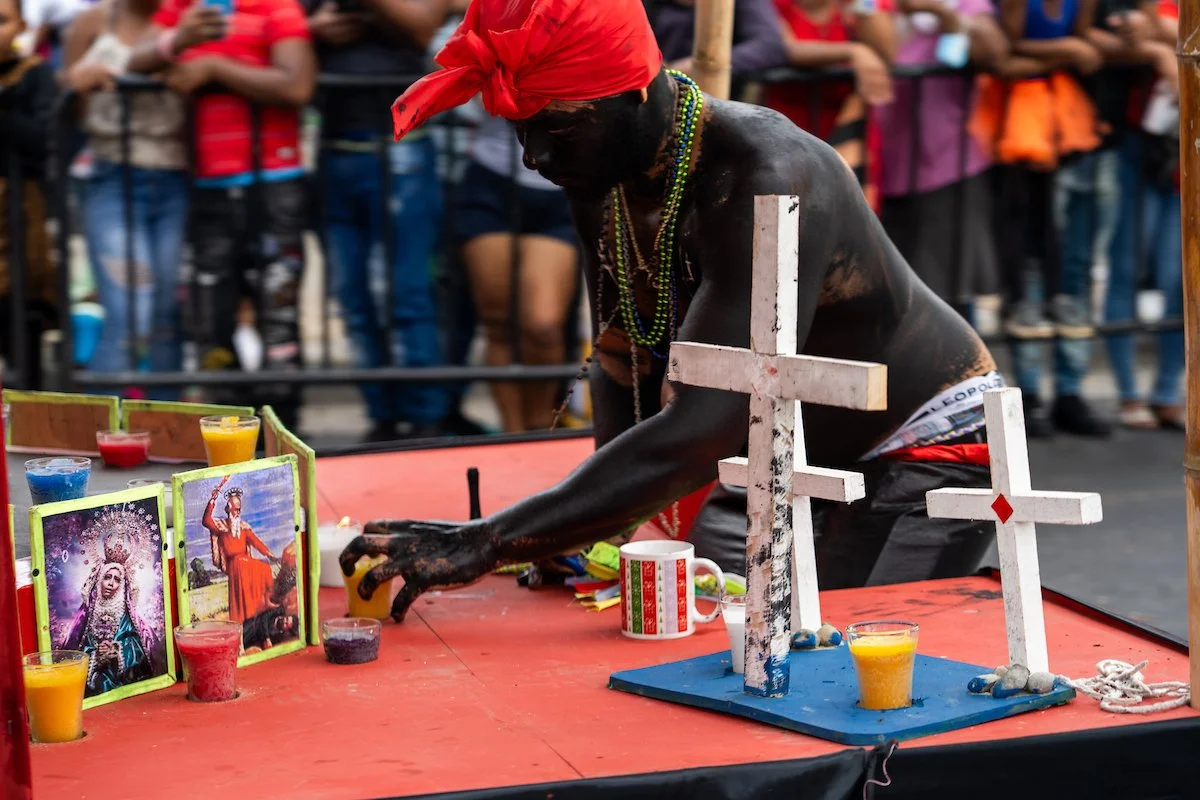 A man with dark skin, wearing a red bandana and multiple necklaces, is performing a ritual at a table with religious icons and candles. He is surrounded by a crowd of onlookers.