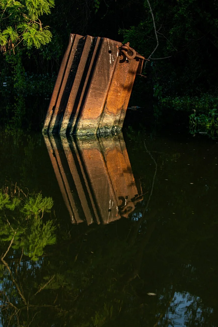 Rusty shipwreck partially submerged in water, surrounded by trees and greenery, with its reflection visible in the water.