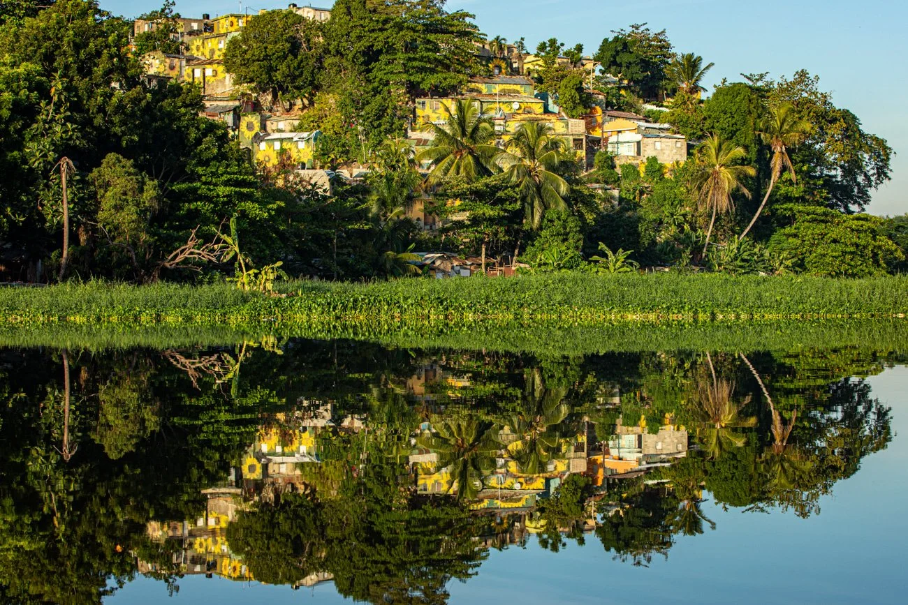 A hillside neighborhood with colorful houses surrounded by lush green trees, reflected in a calm body of water.