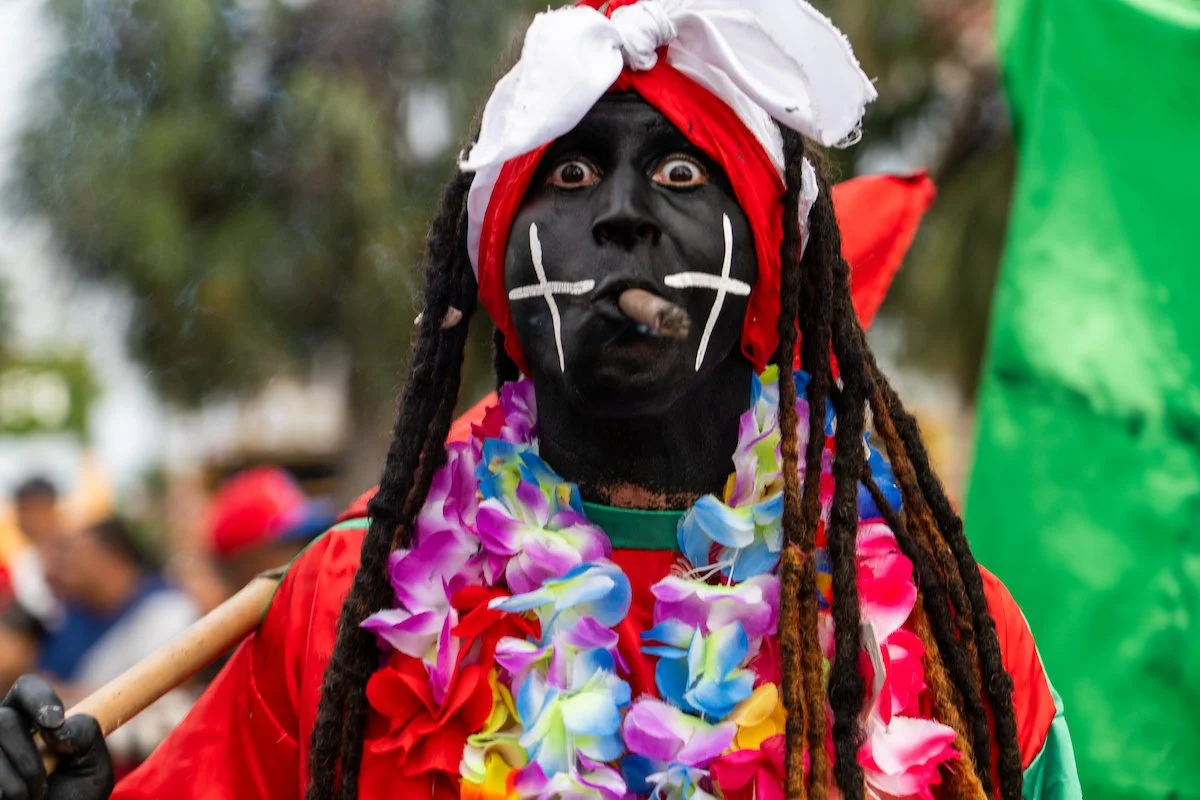 Person dressed in colorful carnival costume with face painted black, white markings, wearing a red bandana and white cloth on head, adorned with a floral lei, holding a stick, and making an expressive face.