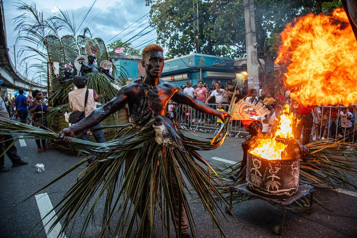 A performer dressed in traditional tribal attire with body paint and palm leaf accessories, dancing next to a burning fire in a street parade with onlookers and colorful floats in the background.
