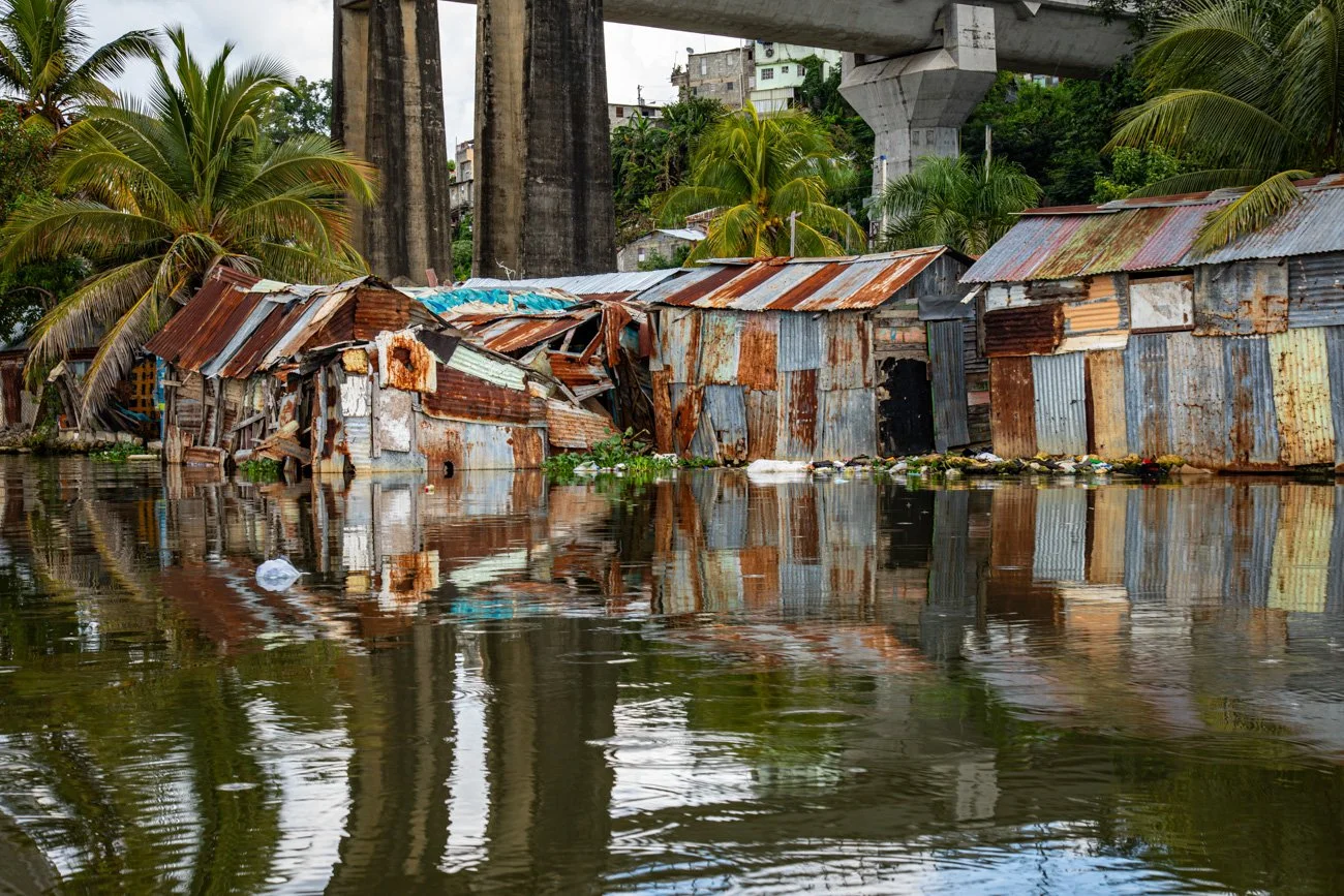 Houses made of rusted metal sheets with palm trees in the background, reflected in flooded water in an urban area with an overpass above.