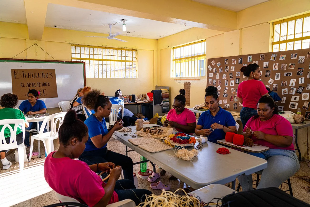 Women sitting at tables in a classroom working on craft projects, with photos and art supplies around.