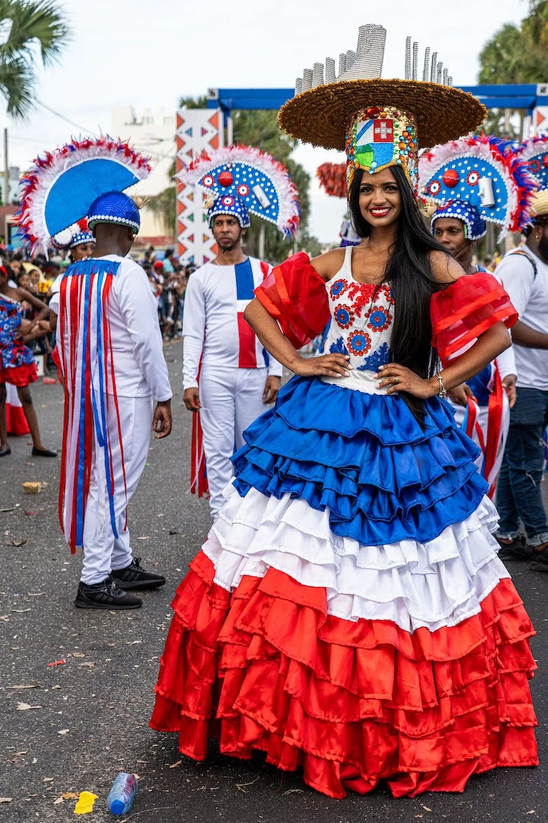 A woman dressed in a colorful, layered dress with red, white, and blue ruffles, wearing a large wide-brimmed hat decorated with a small Colombian flag and flowers, participating in a parade with other people dressed in festive costumes.