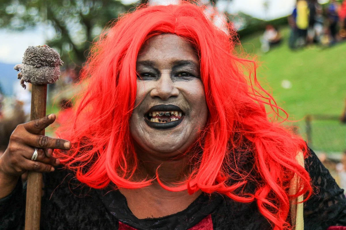 A person with bright red hair, dark makeup, and gold grills on their teeth, holding a stick with a fuzzy object on top, outdoors in a park-like setting.