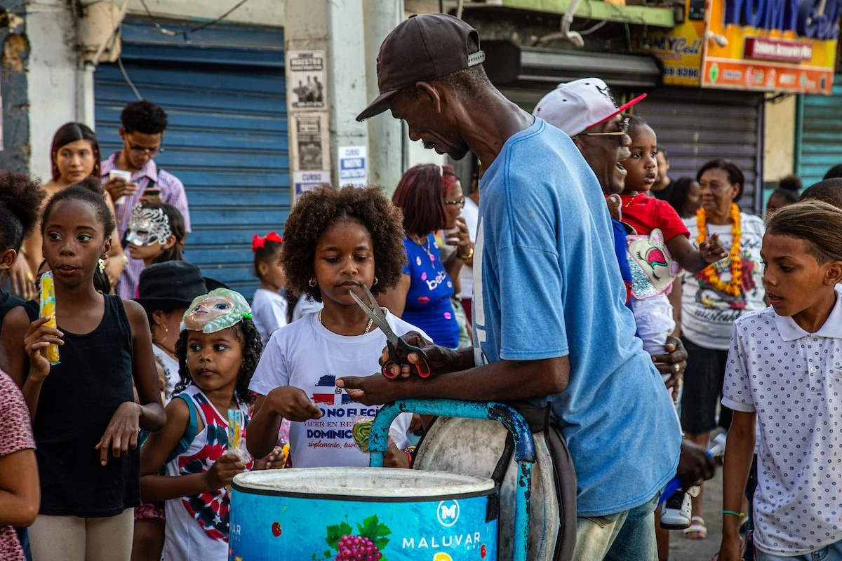 A man cutting a young girl's hair in a busy outdoor market surrounded by children and adults.