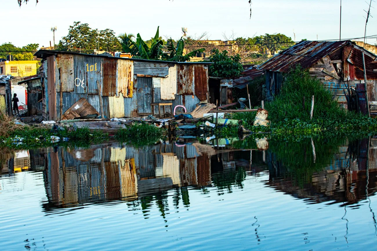 Huts made of rusted metal and wood along a body of water, with reflections of the structures visible in the water, surrounded by greenery.