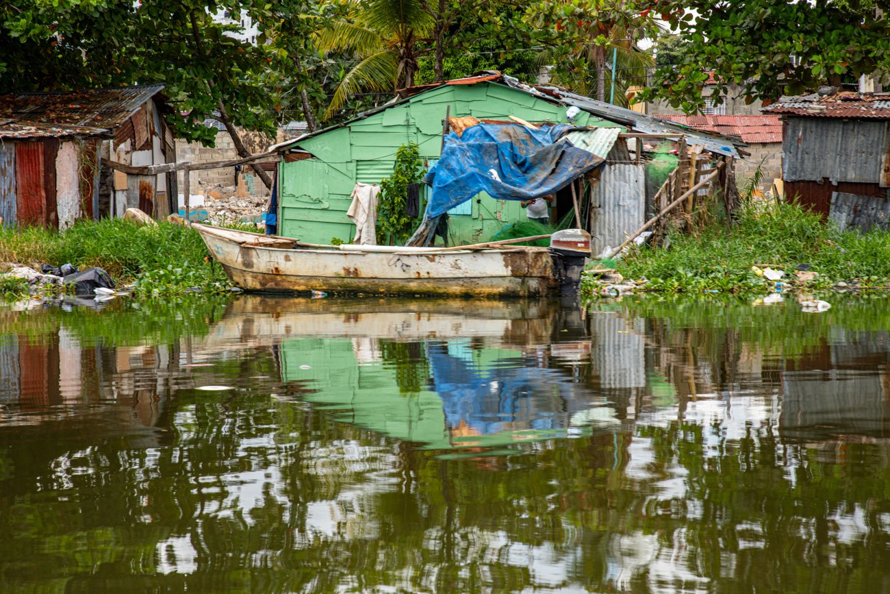 A small, weathered boat is docked in front of a green hut on a waterway, with surrounding makeshift structures and trees reflected in the water.