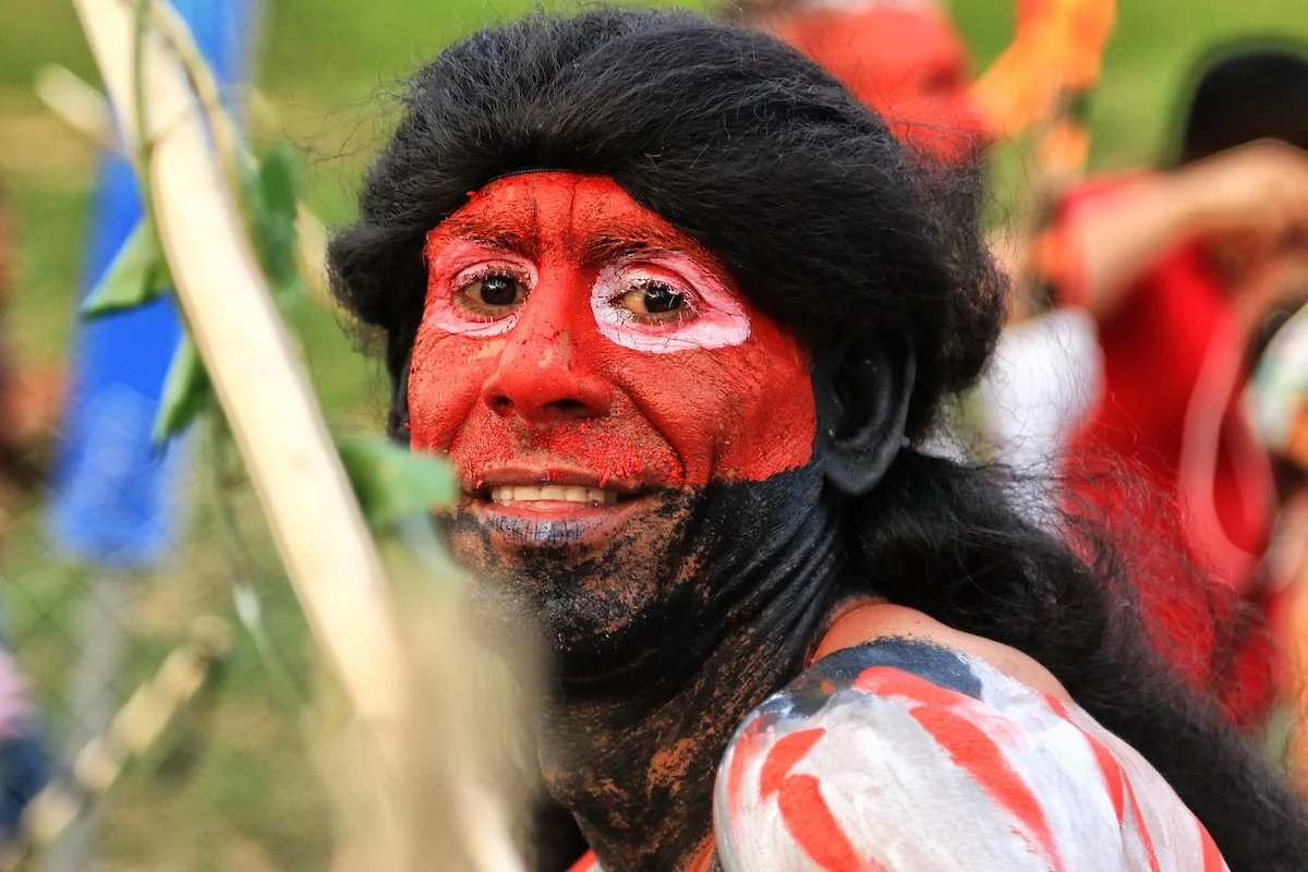 Person with face painted red, black, and white, dressed in traditional attire, performing in a cultural event outdoors.