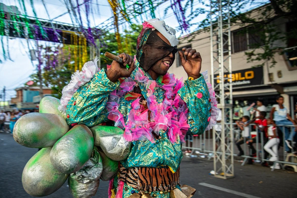 Man dressed in colorful costume with pink flower garland, enjoying a parade with spectators in the background.