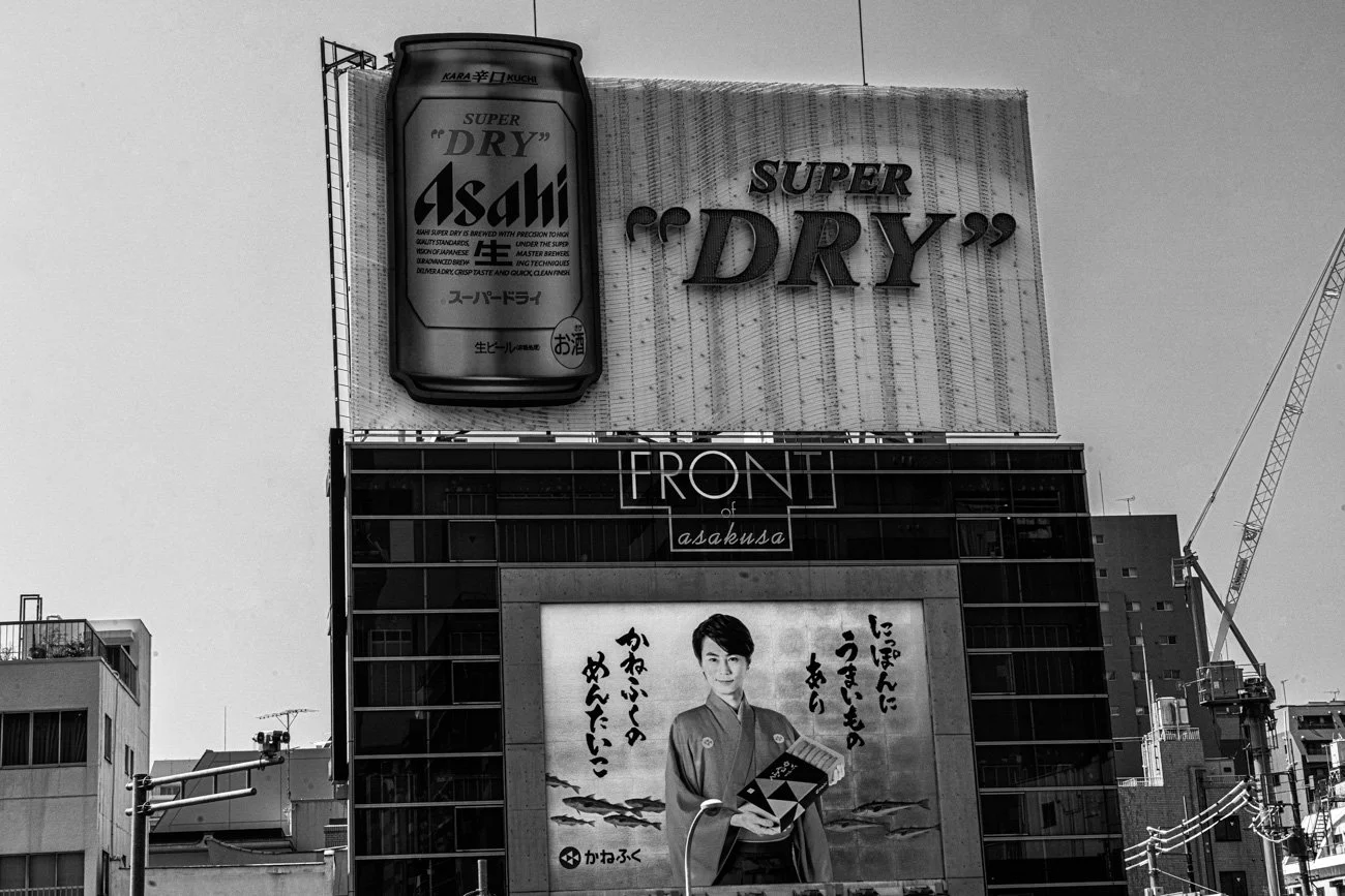 Black-and-white photo of a cityscape with a large billboard advertising Asahi Super Dry beer and a smaller billboard featuring a young man in traditional Japanese clothing holding a book, with Japanese text around him.