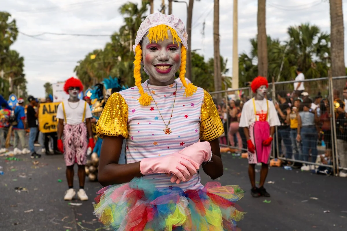 Masquerade parade with people dressed as clowns with colorful costumes, makeup, and rainbow tulle skirts, walking down a street lined with palm trees and spectators.
