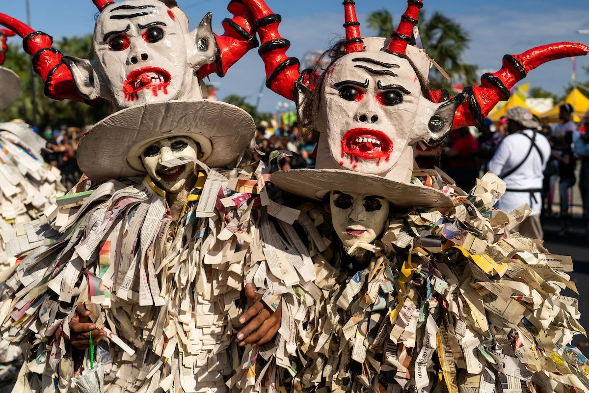 People dressed in elaborate costumes made of paper and wearing large, frightening masks with red horns, black eyes, and blood-like red paint, participating in a festival outdoors with palm trees and yellow tents in the background.