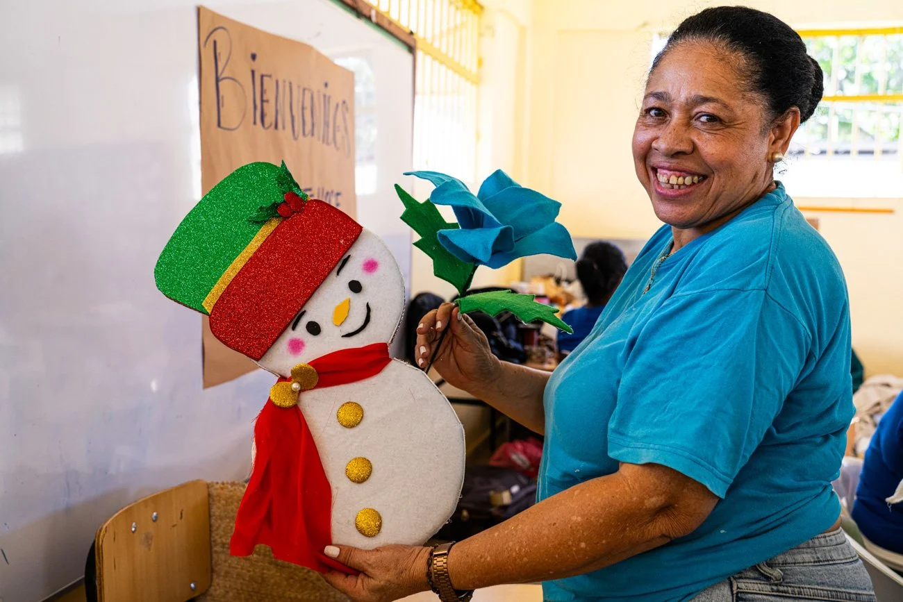 Woman smiling and holding a colorful felt snowman and blue felt flower indoors, with a ‘BIENVENIDAS’ sign in the background.