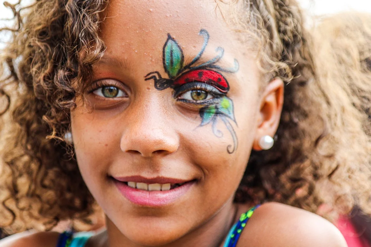 Close-up of a young girl with curly hair, wearing pearl earrings, smiling, with colorful face paint of a ladybug with green and blue leaves on her cheek, and makeup accentuating her eyes.