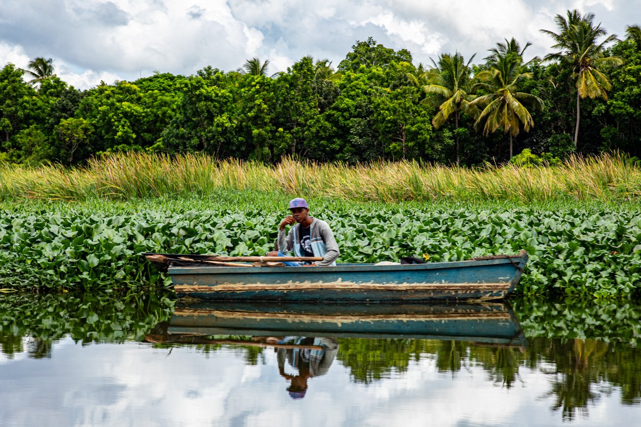 A man in a boat on a calm river surrounded by lush green foliage and trees, including palm trees, with a cloudy sky overhead.