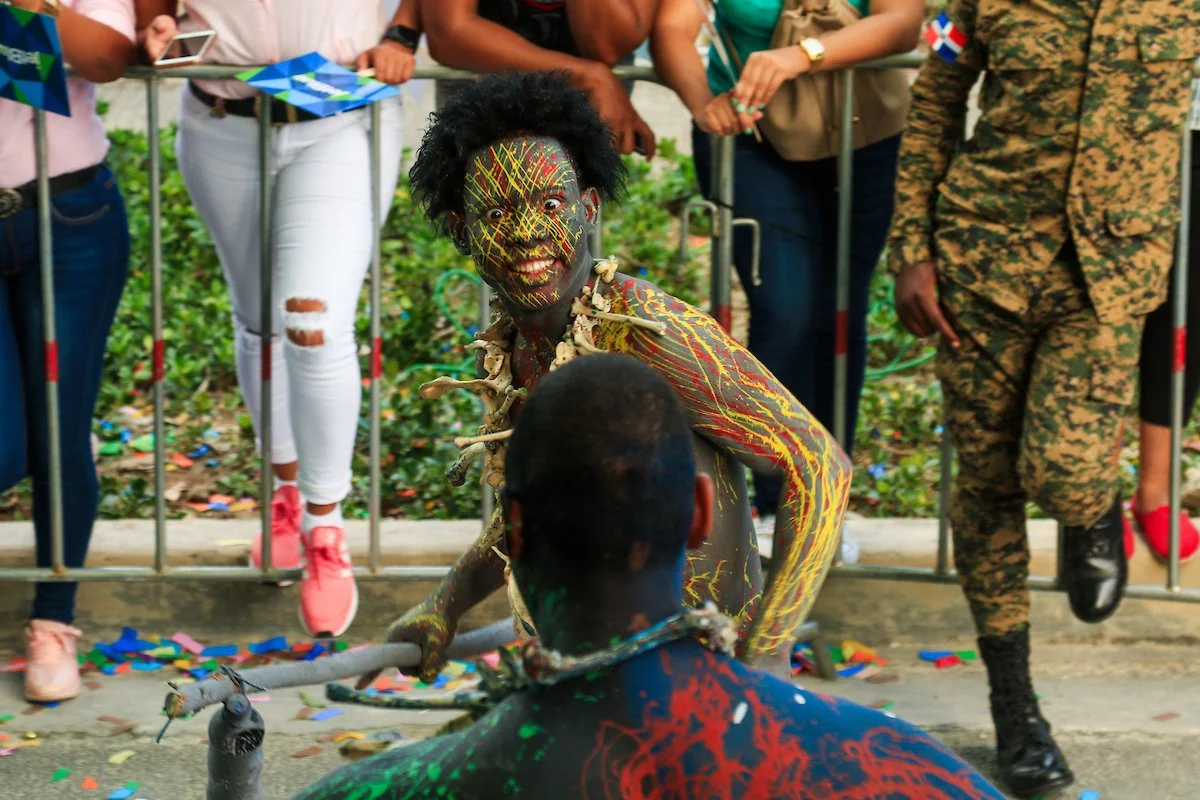 A man with elaborate body paint and costume resembling a tribal or cultural figure, with wild hair and expressive face, engaged with another person at a public event surrounded by onlookers.