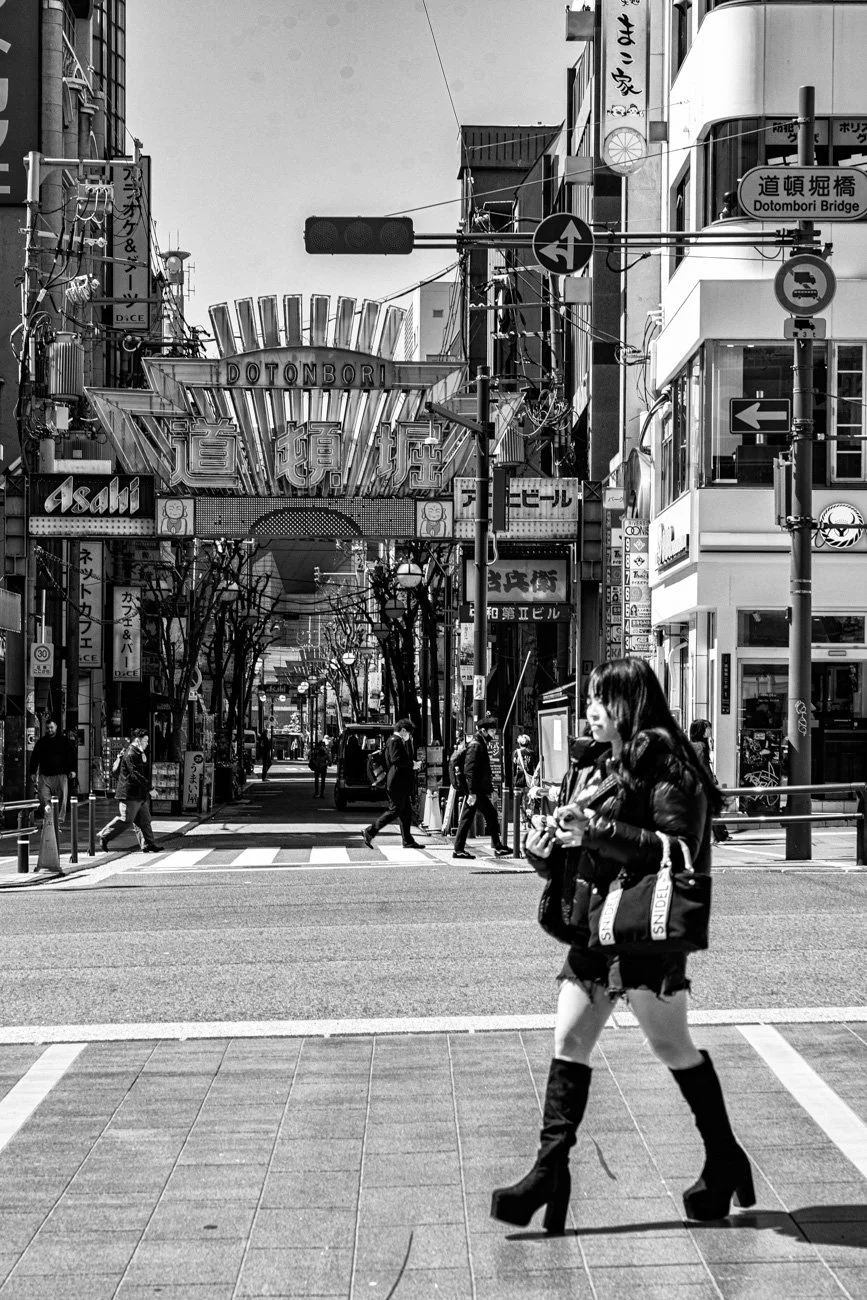 A black and white photo of a busy city street in Japan with people crossing the road and an overhead sign that reads "Dotonbori." There are various storefronts and signs, including Japanese writing and advertisements, with a woman in high-heeled boot