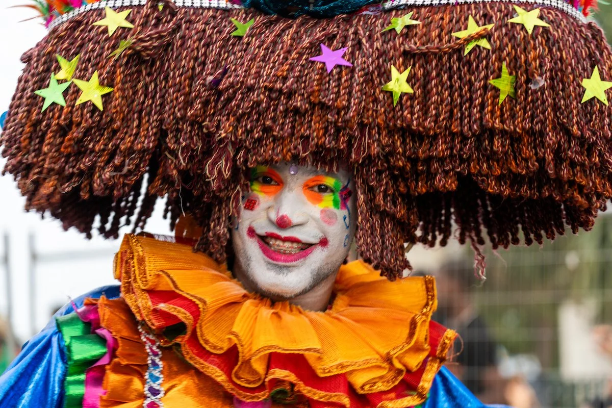 Person dressed as a clown with colorful makeup, wearing a large woven hat decorated with star-shaped stickers, and a ruffled orange collar.