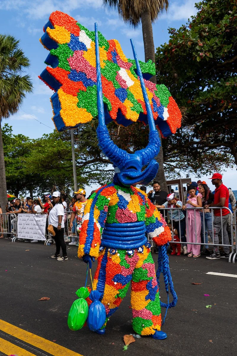 A parade float featuring a large, colorful figure with a blue head and elongated face, dressed in a multicolored suit made of flowers or floral-like decorations, walking along a street lined with spectators. The figure is holding balloons.
