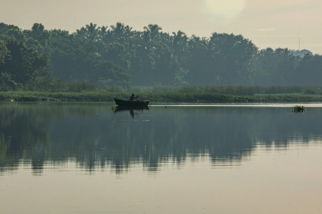 A person in a boat on a calm lake with reflections of trees and greenery, during what appears to be early morning or late afternoon.