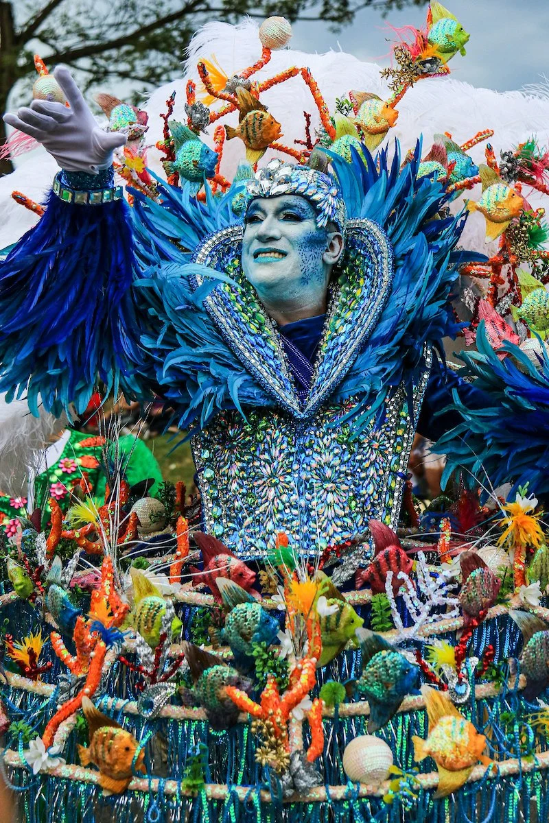 Costumed performer in a vibrant blue and silver aquatic-themed outfit with intricate beadwork, surrounded by decorative sea creatures and coral in a parade or celebration.
