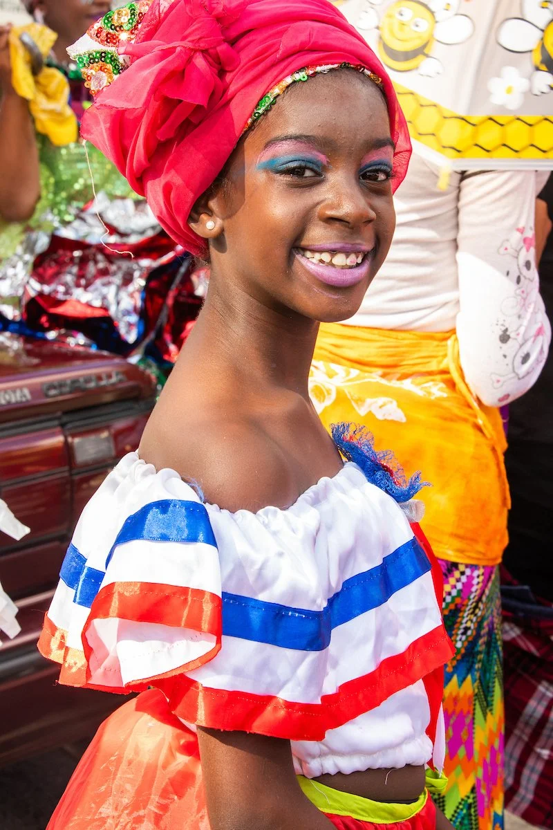 A young woman smiling, wearing colorful eye makeup, a bright red headwrap, and a traditional dress with red, white, and blue stripes, standing among others dressed in traditional clothing.