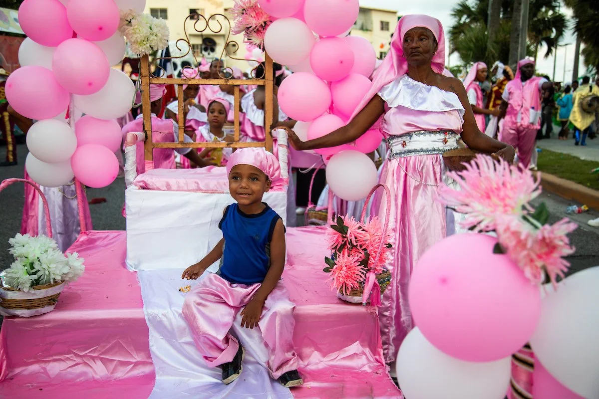 A young boy sitting on a pink decorated float with an elderly woman beside him, surrounded by pink and white balloons and flowers during a parade or celebration.