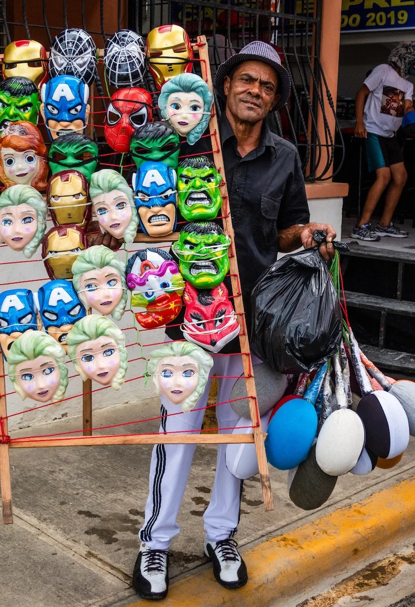Street vendor selling colorful masks resembling characters from popular movies and comics, including superhero masks, princess faces, and animal designs.