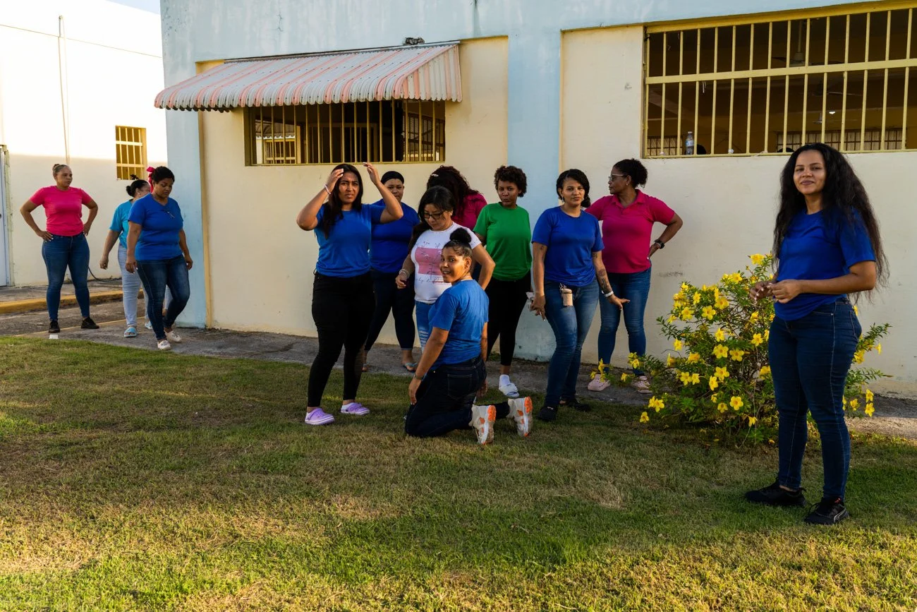 Group of women and girls outdoors near a white building and yellow flowering shrub, some standing and some kneeling, in casual clothing with some wearing blue and pink shirts.