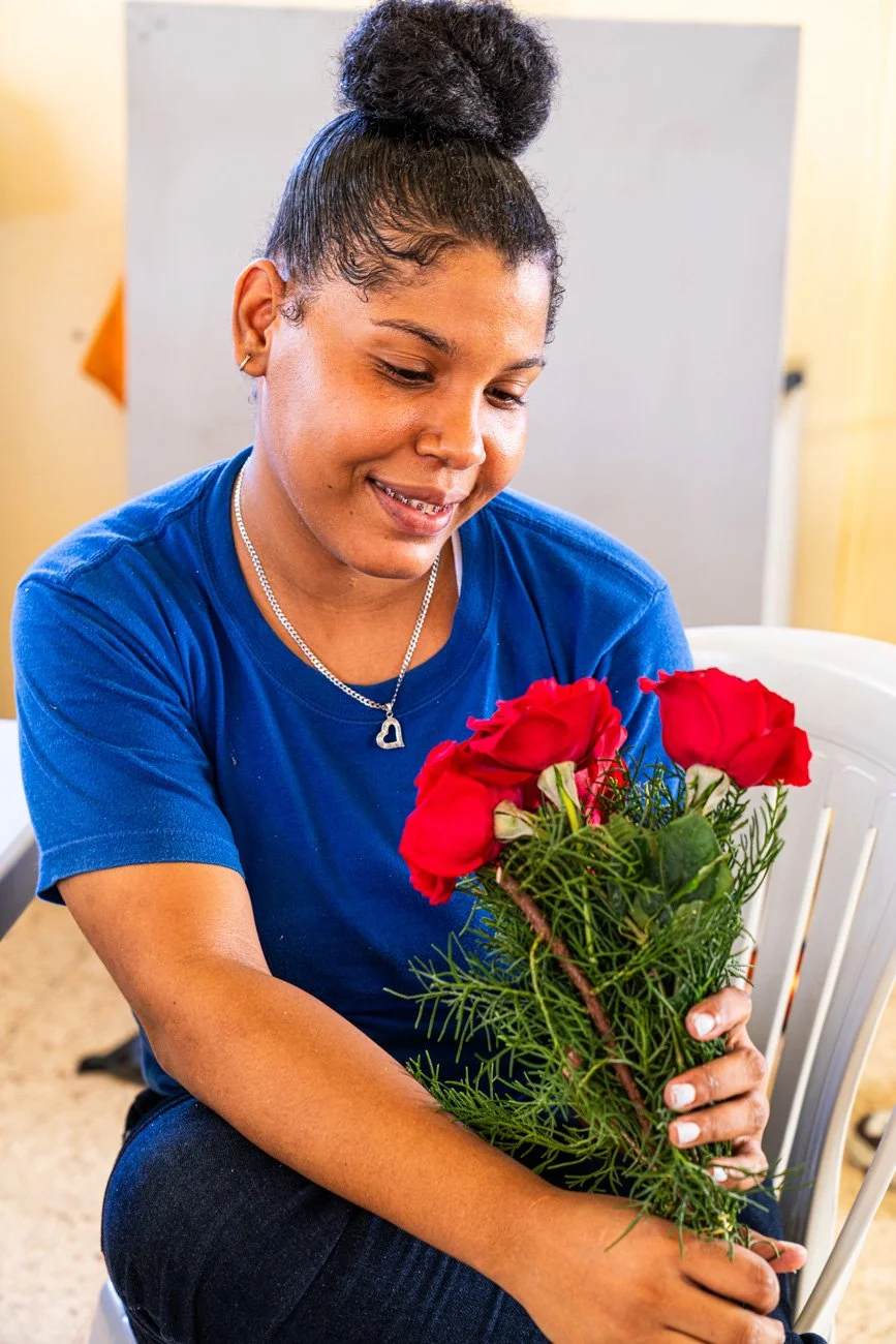 A woman with dark skin and curly hair in a bun, wearing a blue shirt and a necklace with a heart pendant, is sitting and holding a bouquet of red roses and green foliage, smiling gently while looking at the flowers.