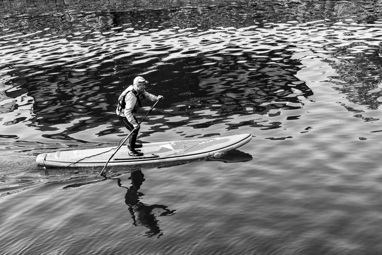 A person stand-up paddleboarding on a calm body of water, wearing a jacket, cap, and sunglasses.