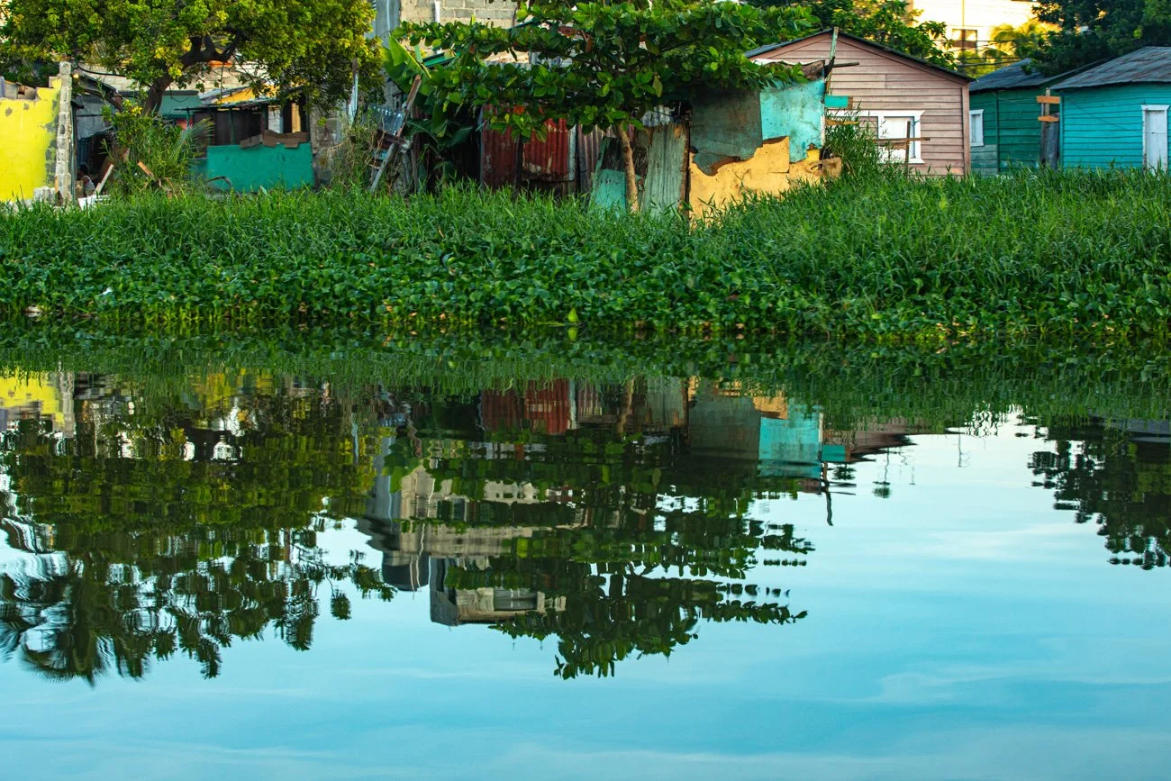 Colorful houses along a canal with lush greenery and trees in the background, reflected in the water.