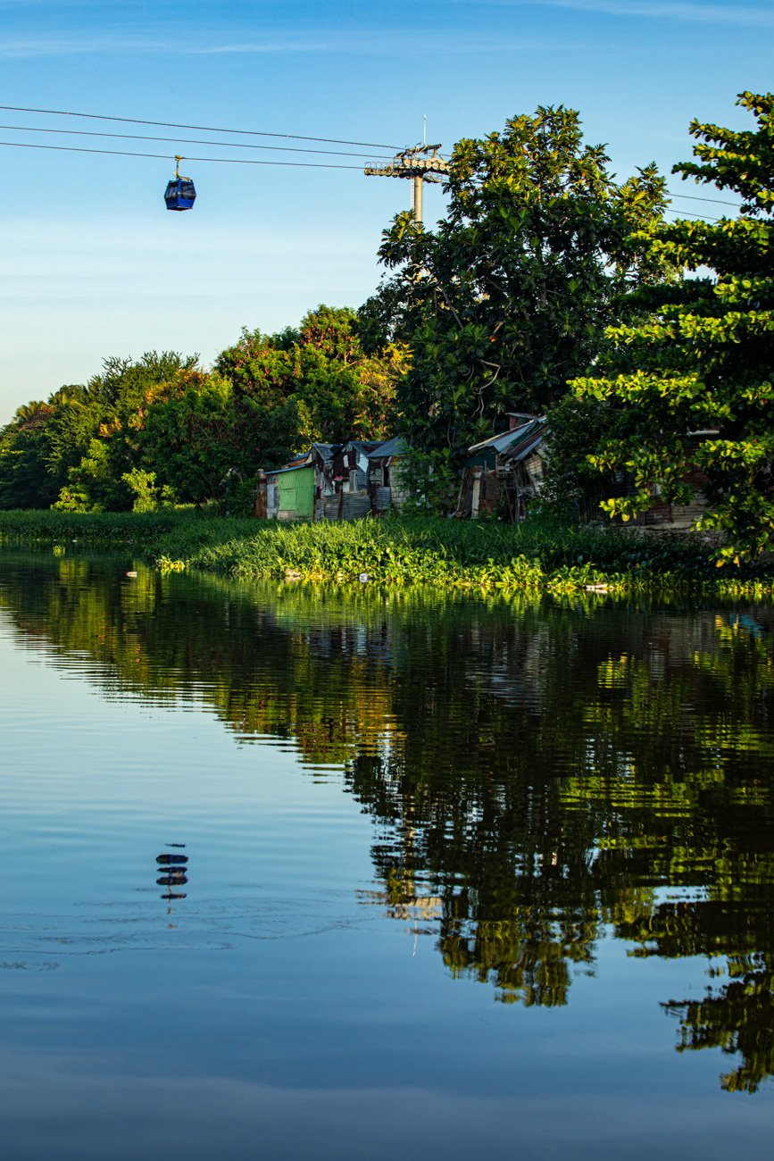 A calm river with green vegetation along the bank, trees, and small houses stacked on the shore. A cable car is seen in the sky, with its reflection in the water, under a clear blue sky.