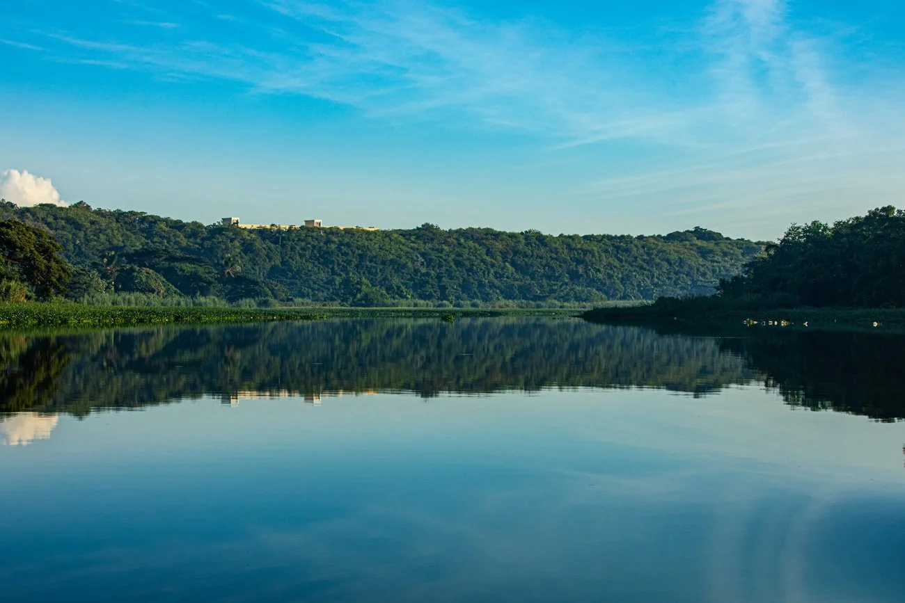 A calm river reflecting a green, forested hillside under a blue sky with wispy clouds.