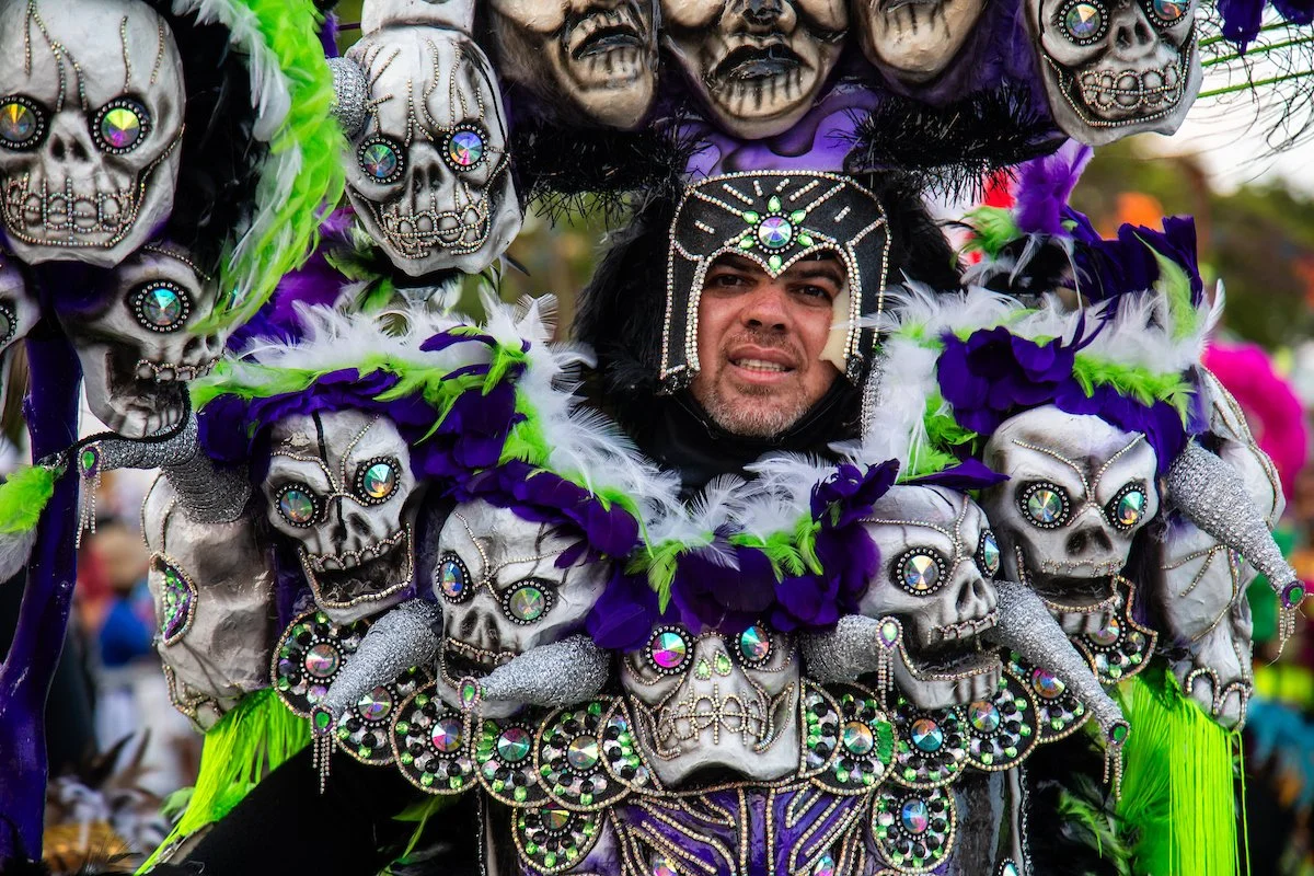 Man dressed in a colorful costume decorated with skulls, feathers, and sparkling ornaments for a Day of the Dead celebration or parade.